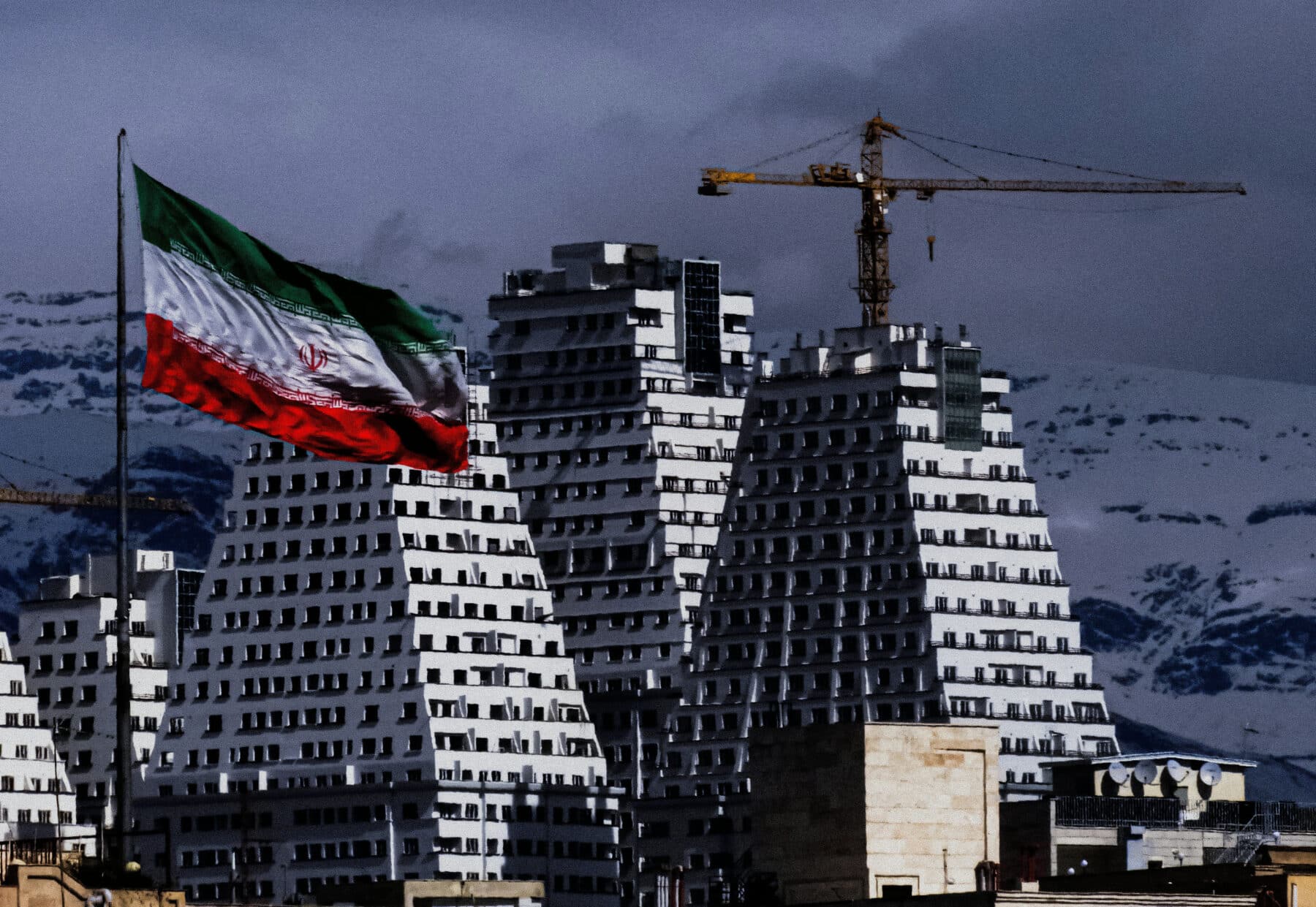 An Iranian flag in the foreground with buildings in Tehran in the background.